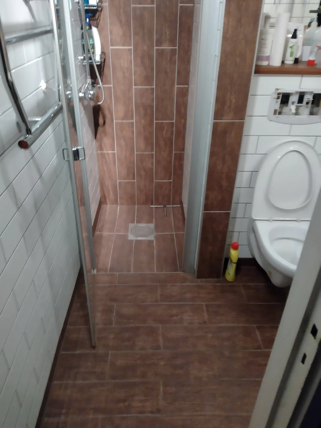 Smalt badrum med brunt träimiterande klinker, duschvägg i glas och vägghängd wc