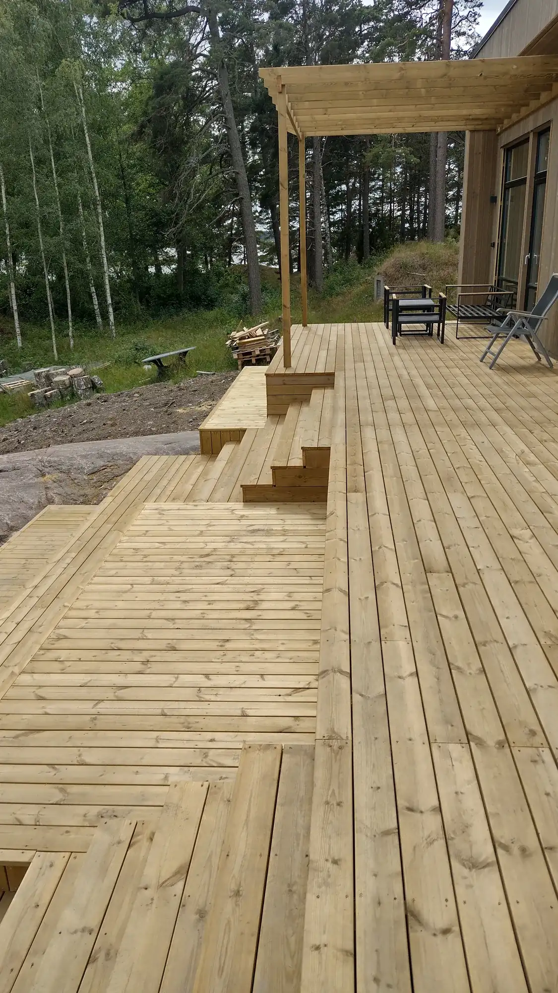 Stort trädäck i flera nivåer med pergola i trä intill ett hus i skogsmiljö