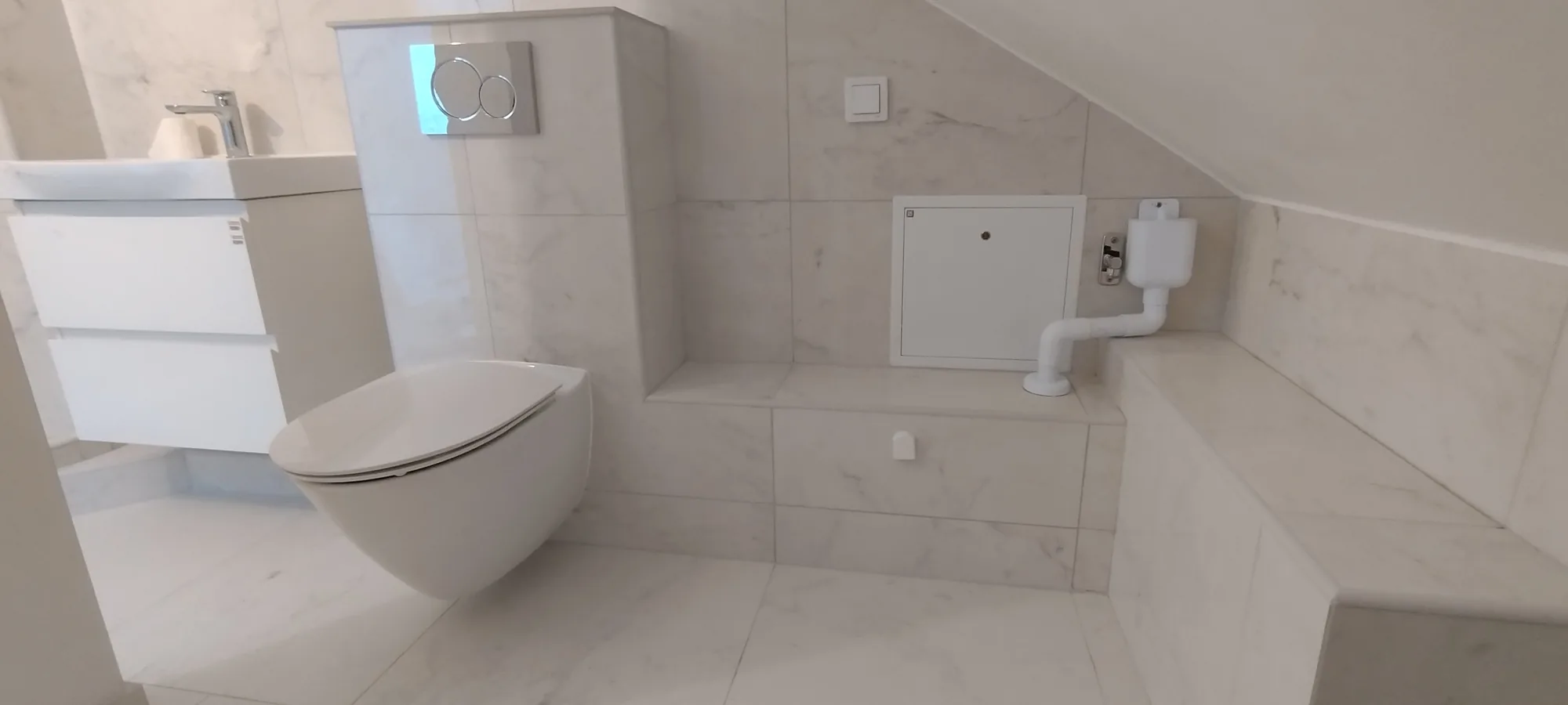 Badrum under snedtak med marmormönstrat kakel, vägghängd wc och inbyggd cistern
